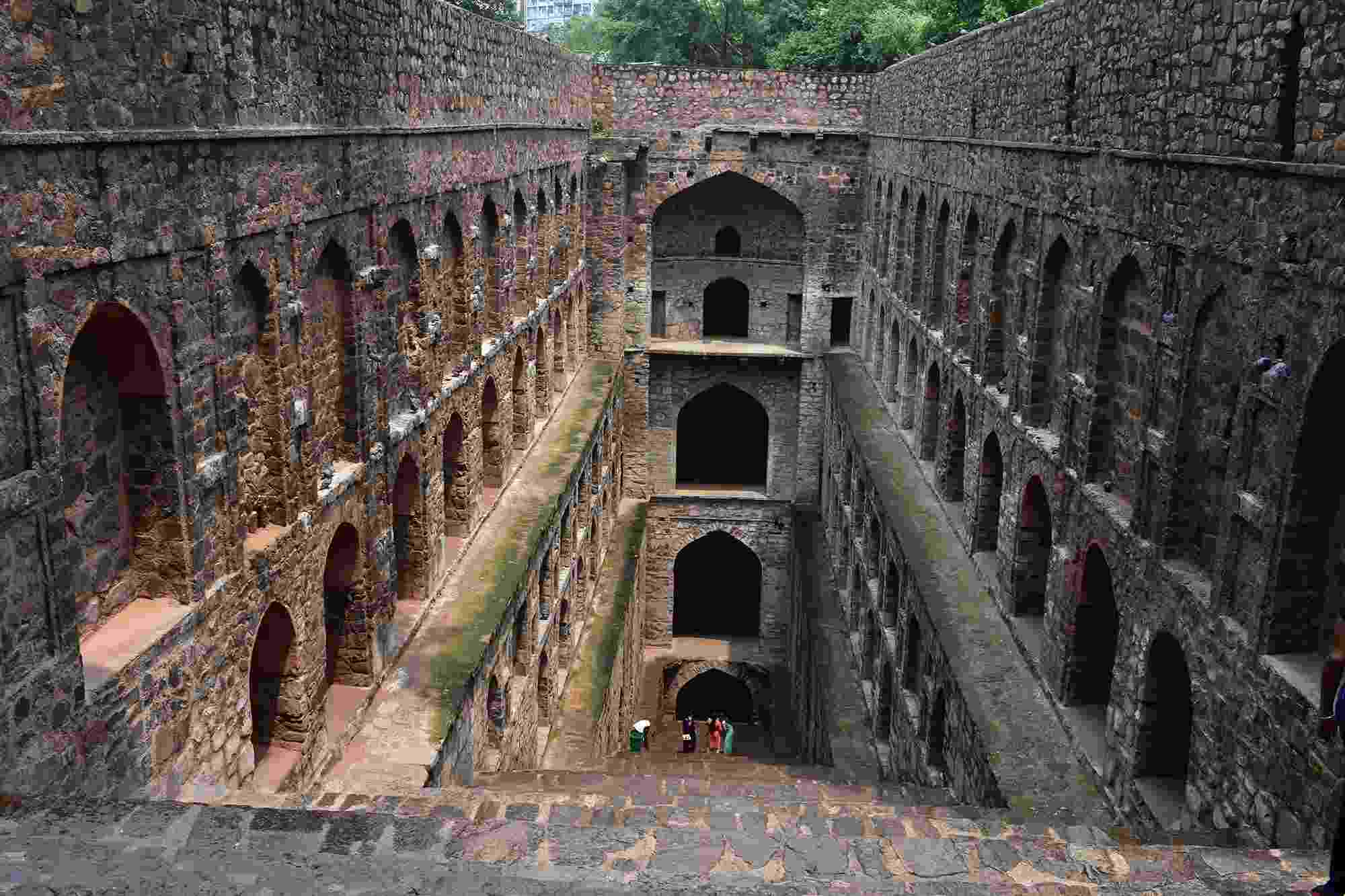 A view down into a historical brown brick stepwell in Delhi. Three levels of the distinctive steps and archways are visible.